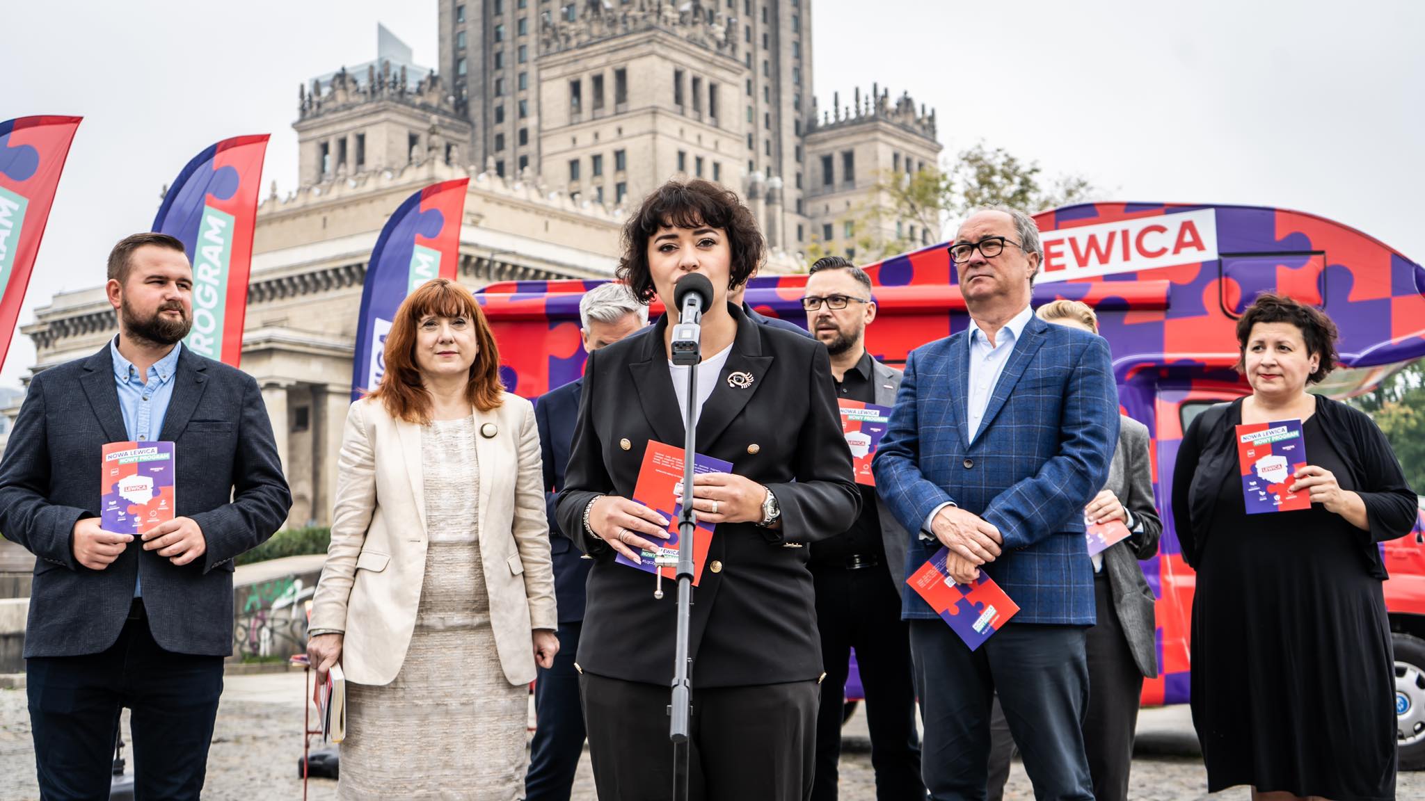 Plenerowa konferencja Nowej Lewicy: kobieta w czarnym garniturze przemawia do mikrofonu, otoczona innymi działaczami trzymającymi ulotki, z kolorowym autobusem promocyjnym i flagami w tle.