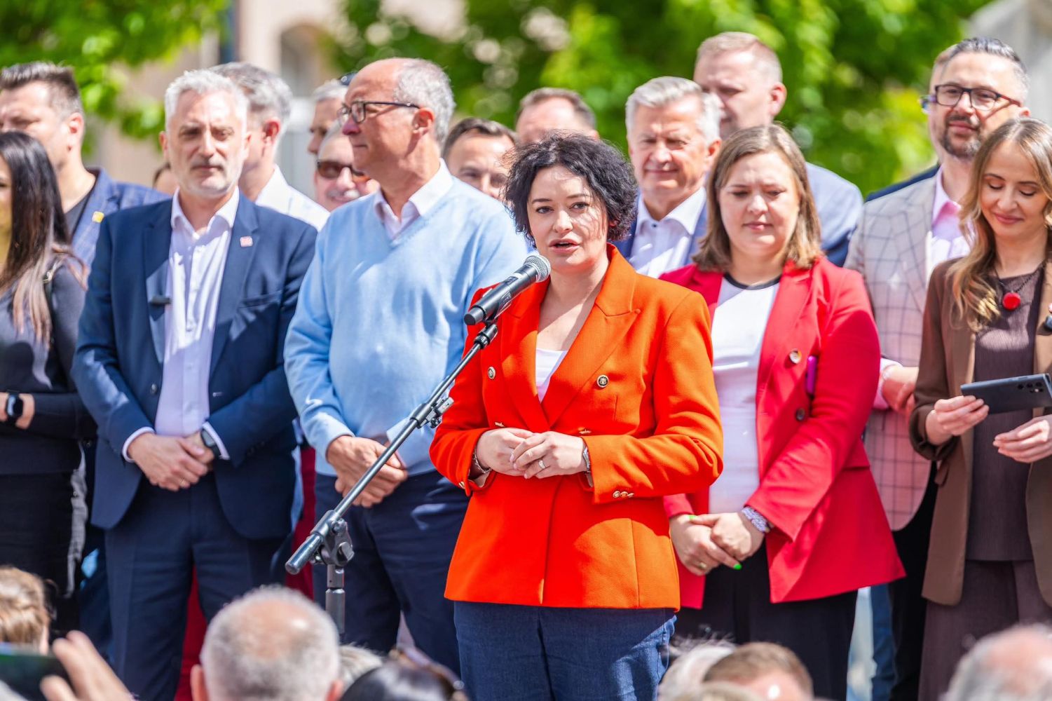 Kobieta w intensywnie pomarańczowej marynarce przemawia do mikrofonu podczas Święta Pracy 2025, otoczona grupą uczestników w garniturach.