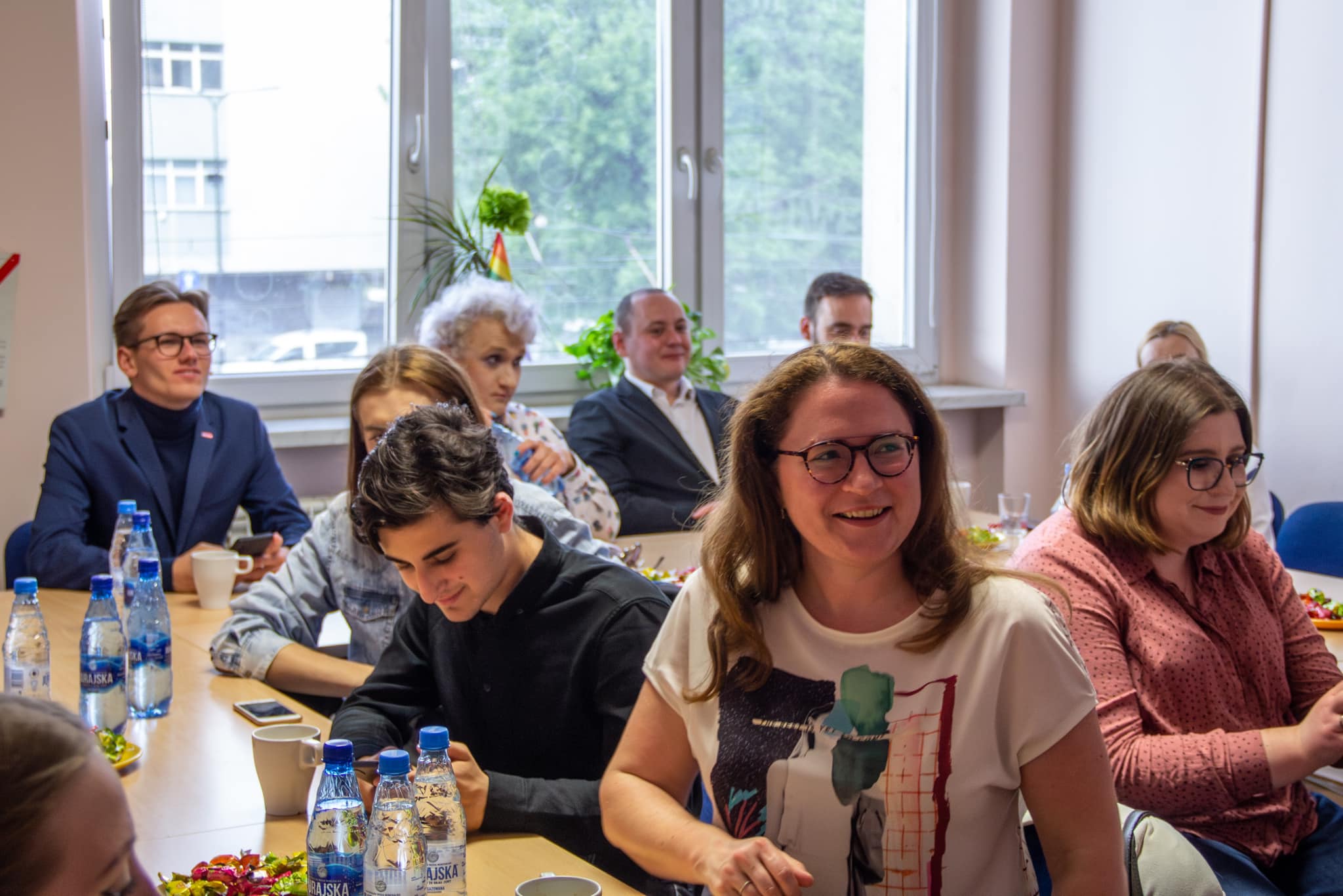 Zdjęcie przedstawia grupę ludzi siedzących przy stole w biurze, z uśmiechającą się kobietą na pierwszym planie oraz innymi uczestnikami spotkania w tle.