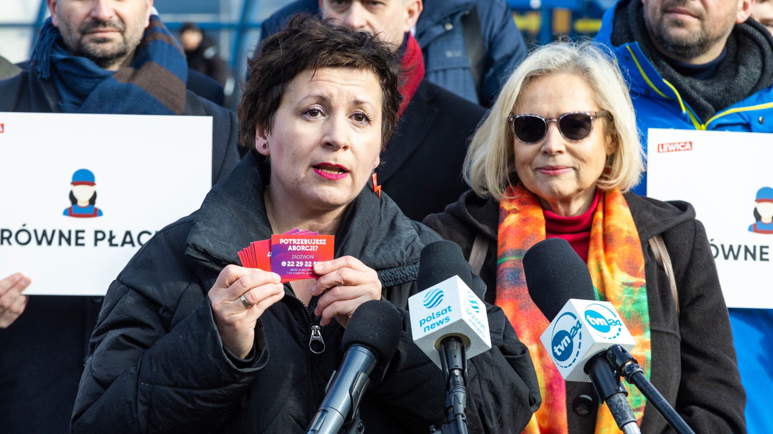Na zdjęciu widoczna jest kobieta mówiąca do mikrofonów, trzymająca ulotki z informacjami, a za nią stoją inni uczestnicy wydarzenia z transparentami.