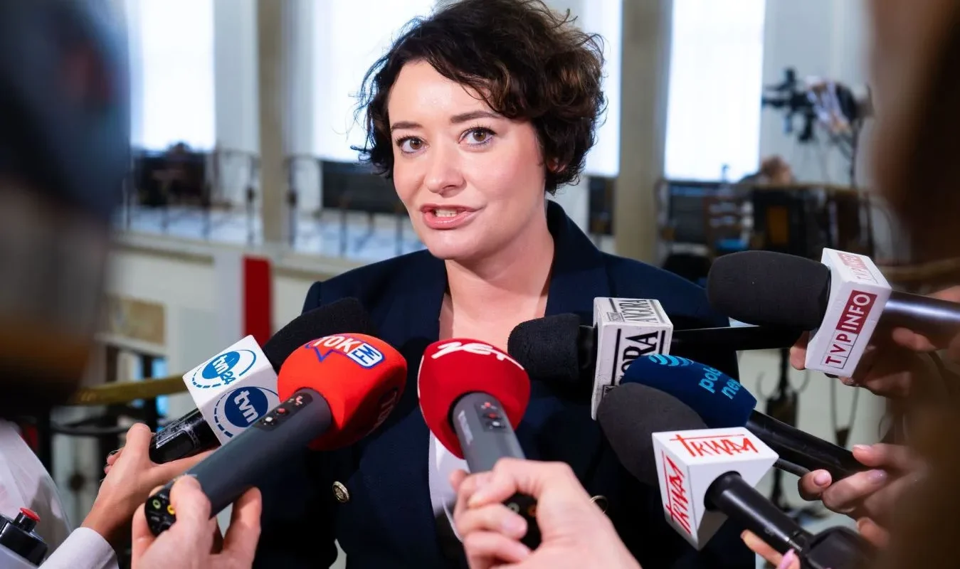 Kobieta odpowiada na pytania dziennikarzy, stojąc wśród licznych mikrofonów podczas konferencji prasowej.