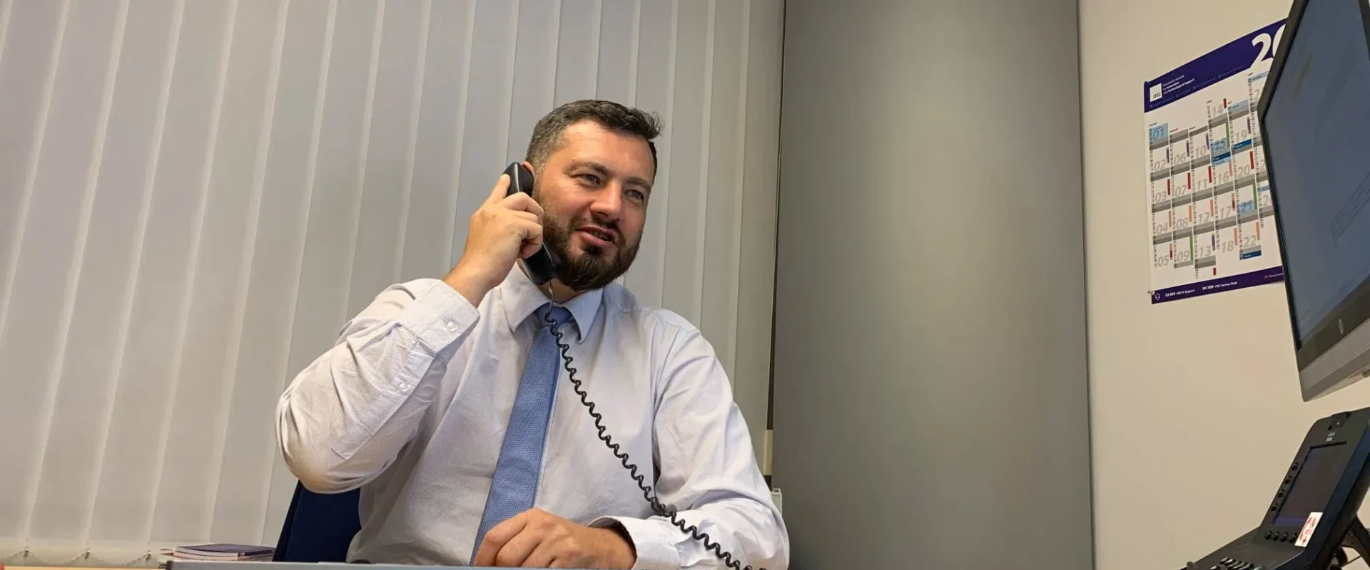 Mężczyzna w białej koszuli i niebieskim krawacie siedzi przy biurku w biurze i rozmawia przez telefon, obok widoczny jest klawiatura, myszka i monitor, a na ścianie kalendarz.