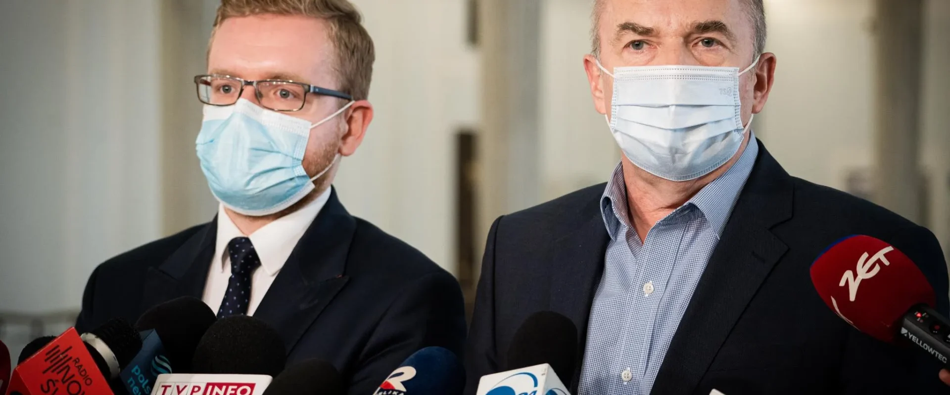 Na zdjęciu widać dwóch mężczyzn w maseczkach, stojących przed mikrofonami i udzielających wywiadu.