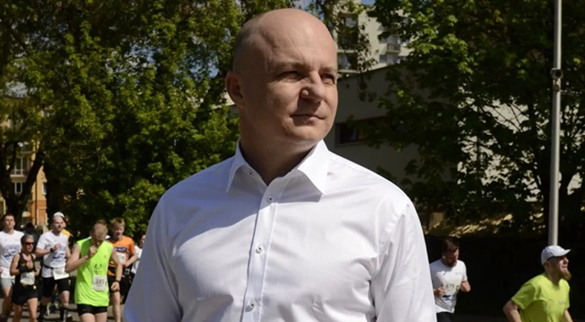 Mężczyzna w białej koszuli stoi na tle grupy biegaczy podczas wydarzenia sportowego na zewnątrz, wśród drzew.
