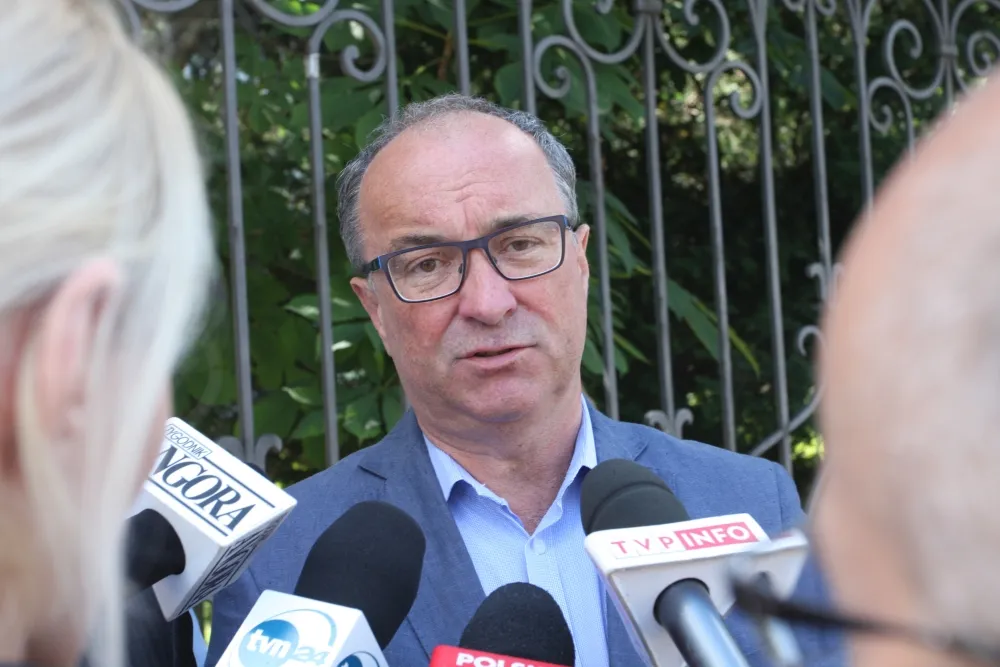 Mężczyzna w okularach, granatowym garniturze i jasnoniebieskiej koszuli przemawia do mikrofonów dziennikarzy na zewnątrz, na tle metalowego ogrodzenia i zieleni.