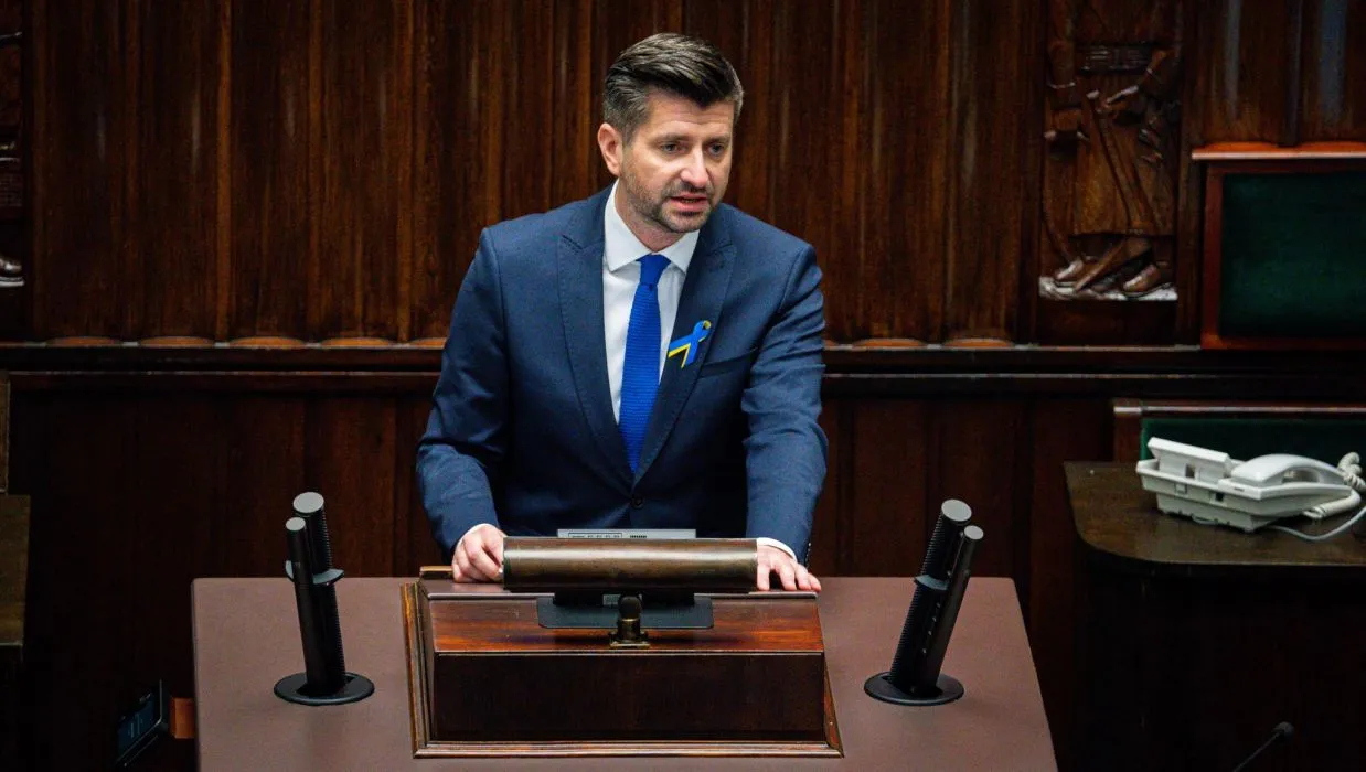 Mężczyzna w garniturze przemawia w sali obrad, stojąc przed mównicą.