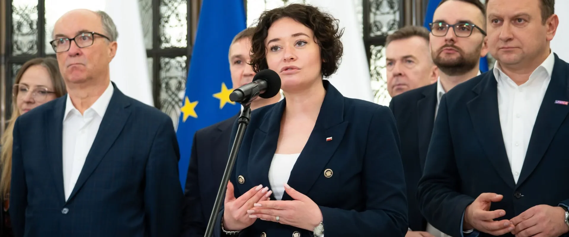 Polityczka Lewicy przemawia do mikrofonu podczas konferencji prasowej, w otoczeniu innych działaczy i flag Unii Europejskiej.