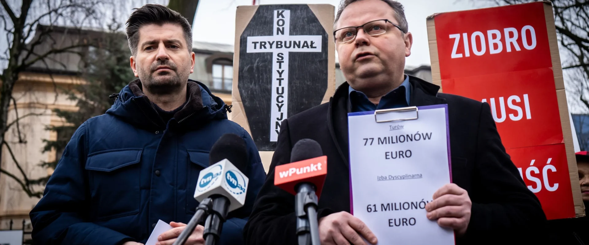 Dwóch mężczyzn podczas konferencji prasowej stoi przy mikrofonach, w tle widoczne są plakaty i banery związane z Europejską Konwencją Praw Człowieka, w tym z napisem Ziobro oraz kartką z informacją o 77 milionach euro.