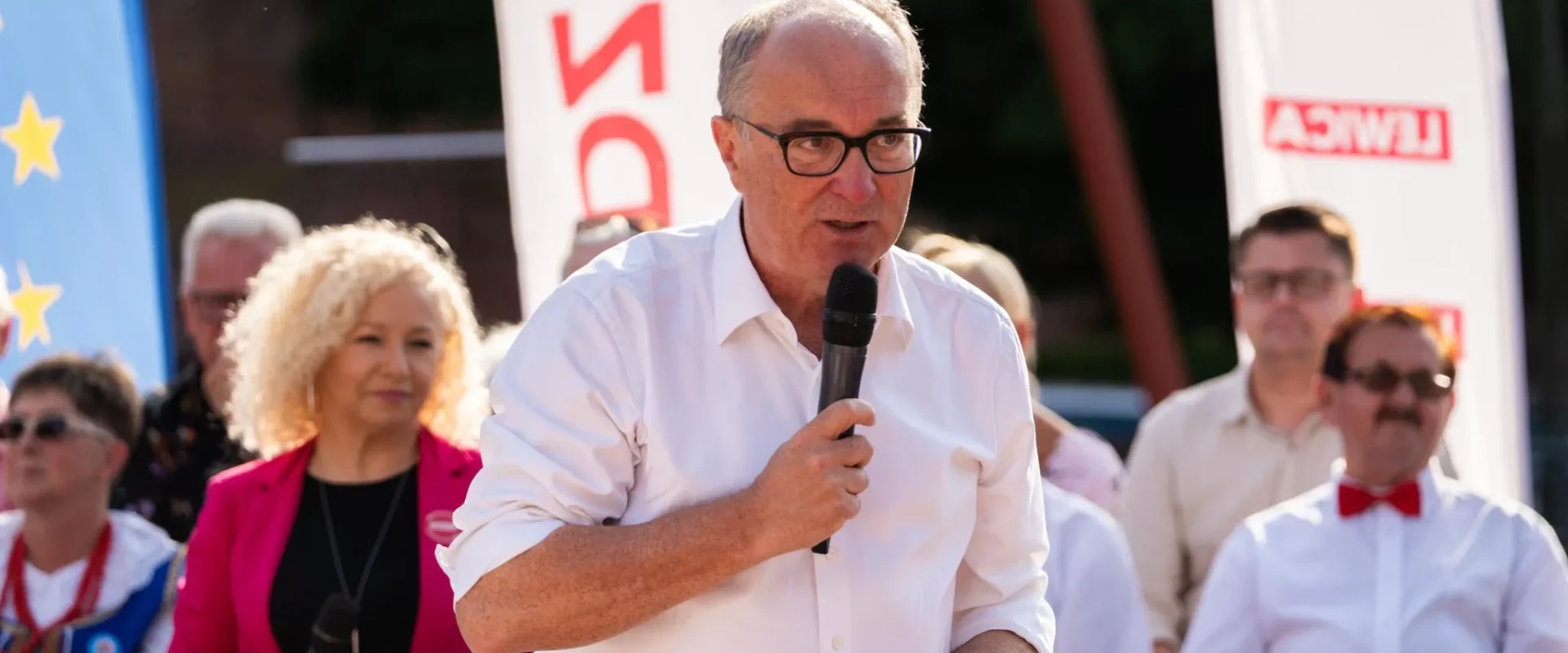 Mężczyzna w białej koszuli i czarnych okularach przemawia do zgromadzonych, trzymając mikrofon, na tle bannerów i publiczności.