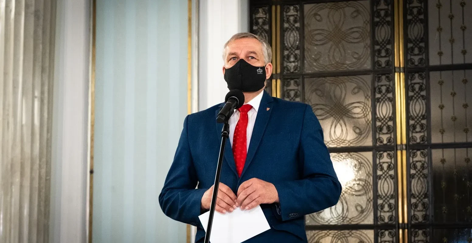 Mężczyzna w granatowym garniturze i czerwonym krawacie stoi przy mikrofonie, trzyma kartki i ma czarną maseczkę na twarzy, w tle ozdobne drzwi.