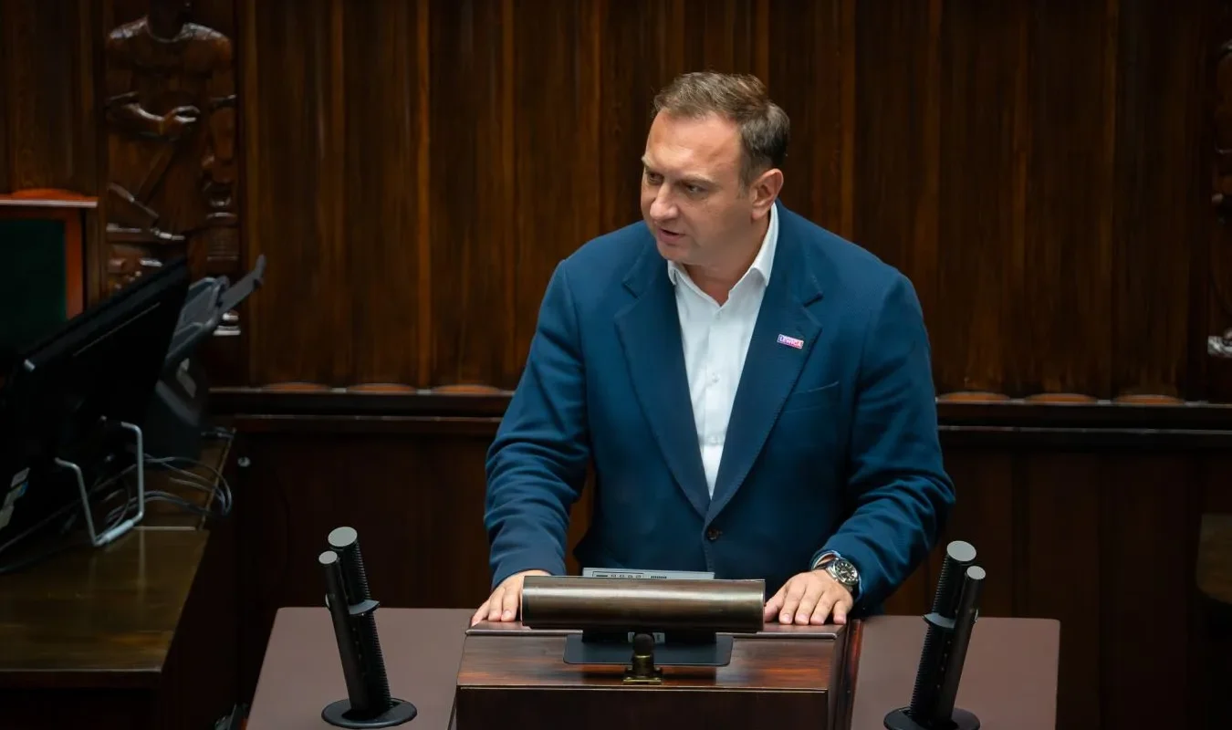 Mężczyzna w niebieskim garniturze stoi przy mównicy w sali plenarnej, otoczony mikrofonami.