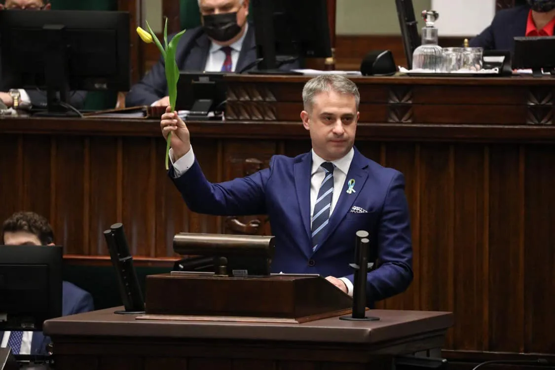 Mężczyzna w garniturze trzyma zieloną roślinę w podniesionej ręce, stojąc za biurkiem w sali obrad.
