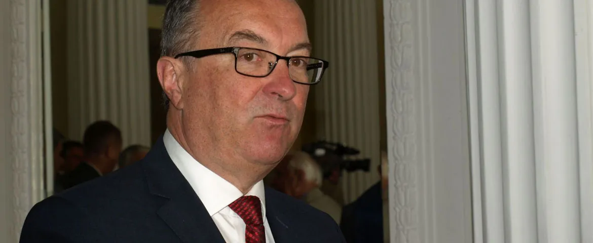 Mężczyzna w garniturze, czarnych okularach i czerwonym krawacie stoi przy wejściu w eleganckim wnętrzu.