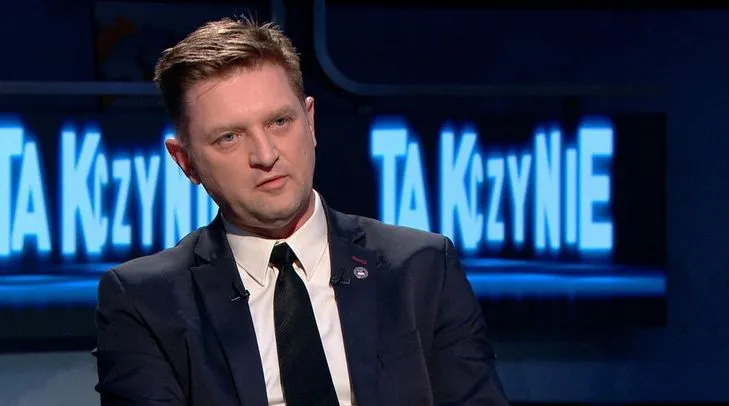 Mężczyzna w ciemnym garniturze i czarnym krawacie siedzi w studiu telewizyjnym, a w tle widoczne są niebieskie napisy.