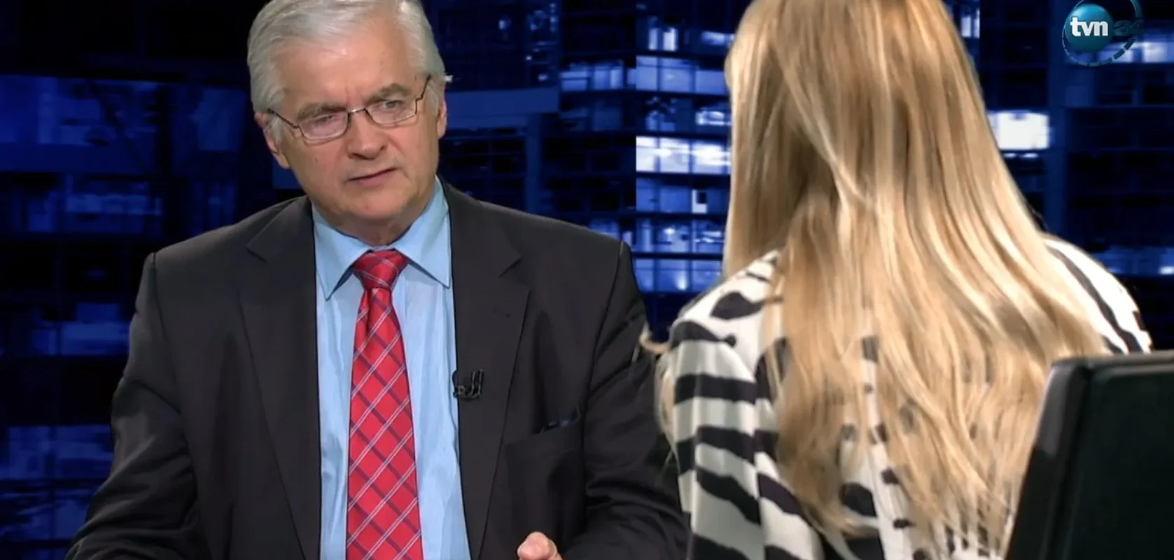 Mężczyzna w garniturze z czerwonym krawatem rozmawia z kobietą o blond włosach w studiu telewizyjnym, z logo TVN w tle.