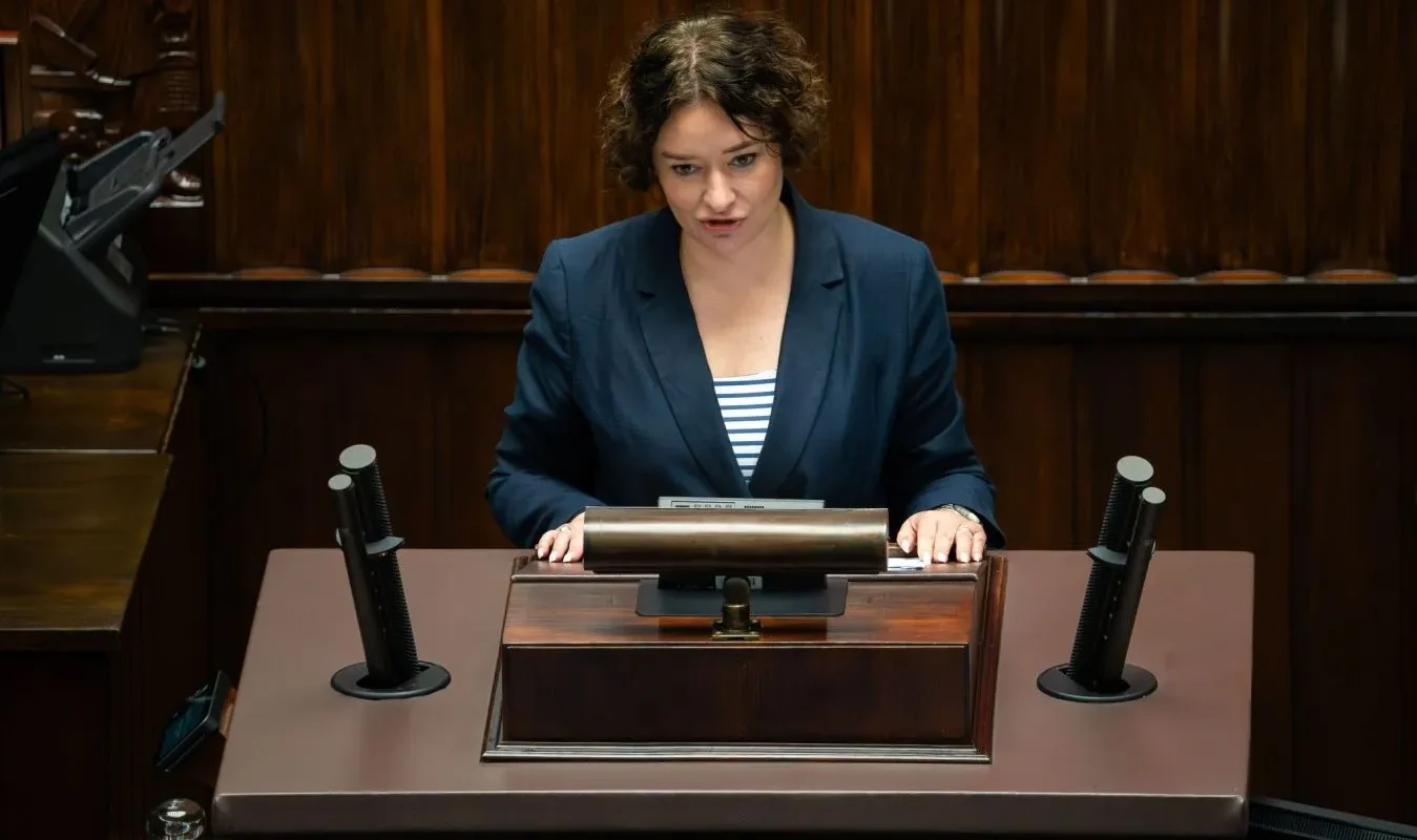 Kobieta w granatowej marynarce stoi za drewnianym pulpitem w sali obrad, po obu stronach widoczne są mikrofony.