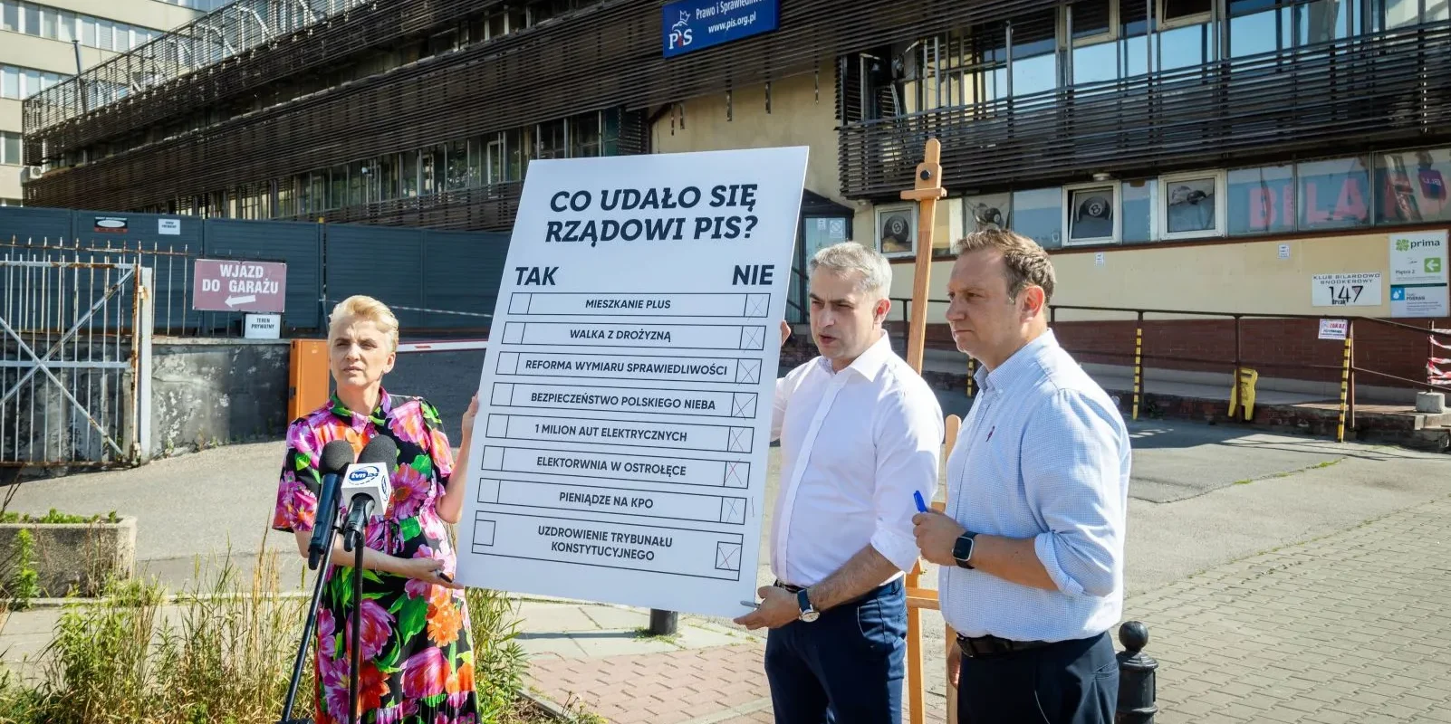 Trzy osoby stoją na zewnątrz obok dużej tablicy z pytaniem „Co udało się rządowi PiS?”, na której widnieje lista obietnic do oceny podczas konferencji prasowej.