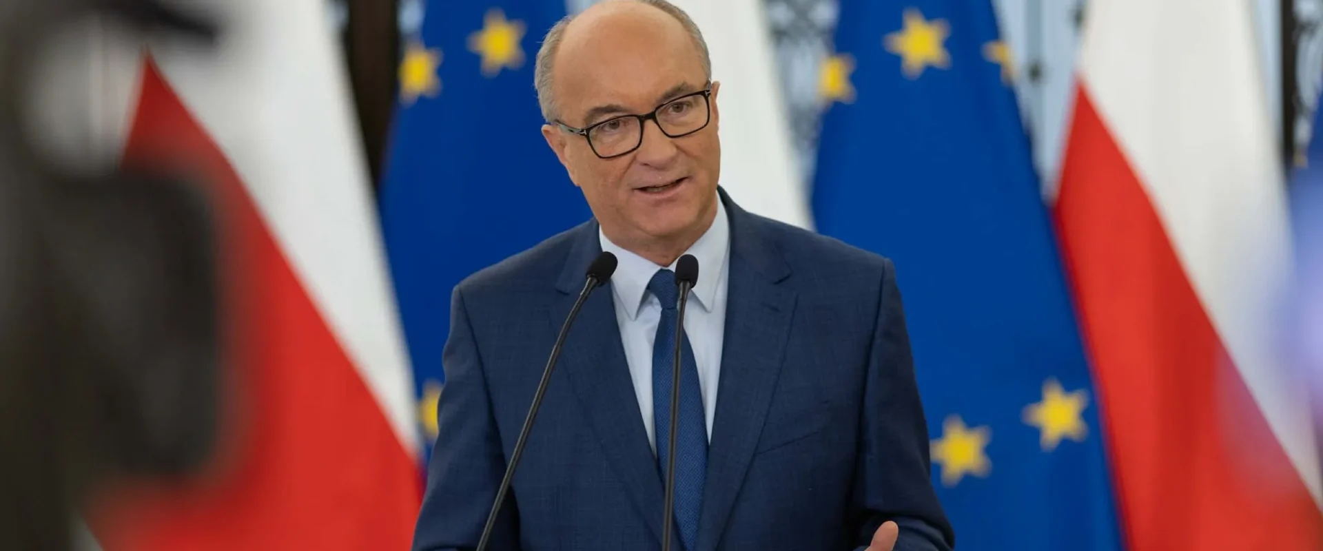 Mężczyzna w garniturze przemawia do mikrofonów podczas konferencji prasowej, na tle widoczne flagi Polski i Unii Europejskiej.