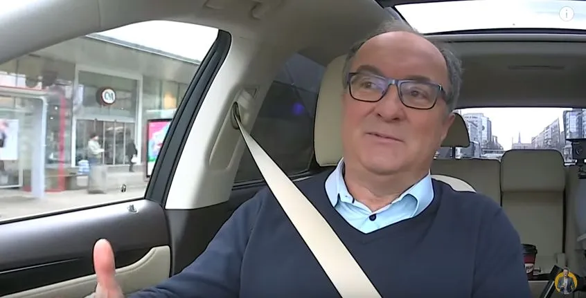Mężczyzna w okularach siedzi na przednim siedzeniu samochodu z zapiętym pasem, gestykulując dłonią, a przez szybę widać zabudowaną ulicę.