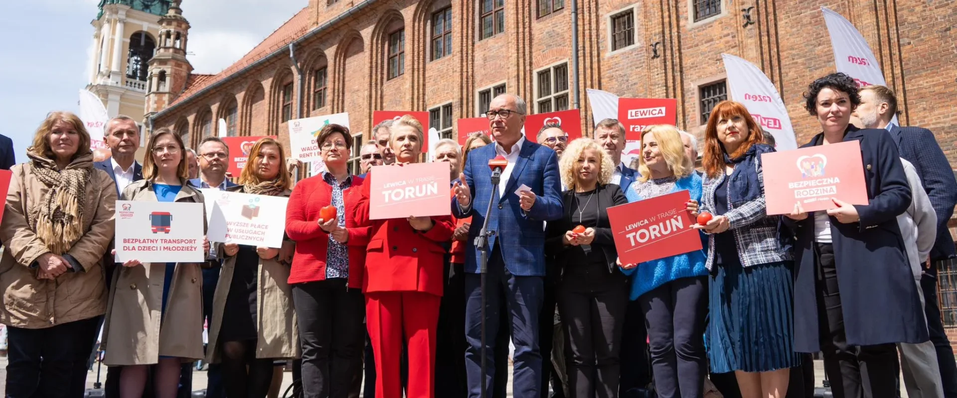 Grupa osób na placu przed historycznym budynkiem trzyma plakaty Lewicy z napisem Toruń, a na środku mężczyzna w błękitnym garniturze przemawia do mikrofonu.