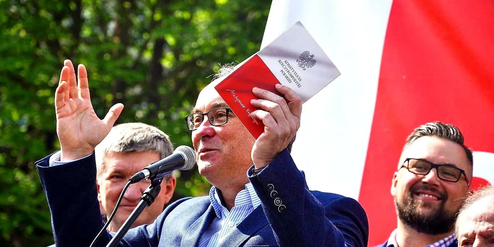 Mężczyzna w granatowym garniturze przemawia do mikrofonu, trzymając w dłoni egzemplarz Konstytucji RP podczas zgromadzenia z biało‑czerwoną flagą w tle.