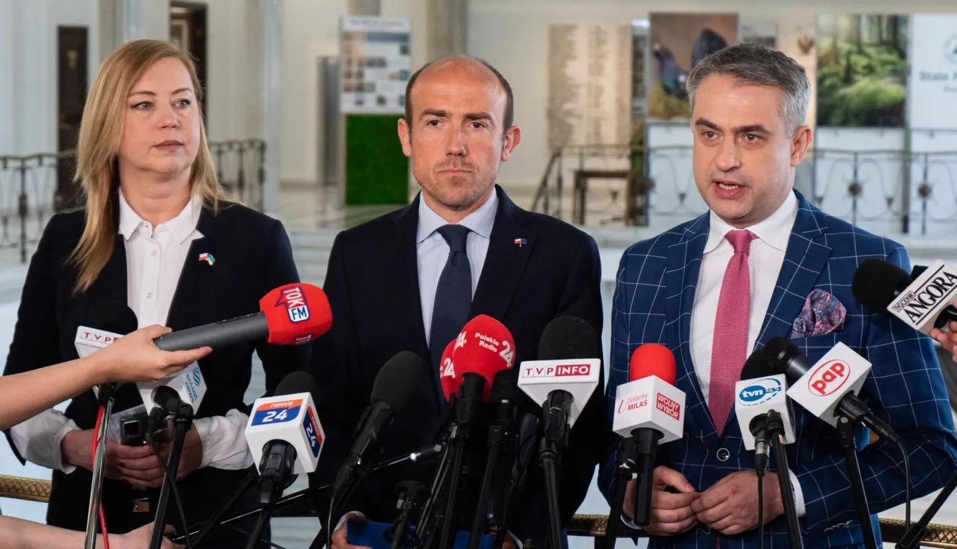 Trzech polityków stoi podczas konferencji prasowej, otoczonych licznymi mikrofonami reporterów w wnętrzu budynku.