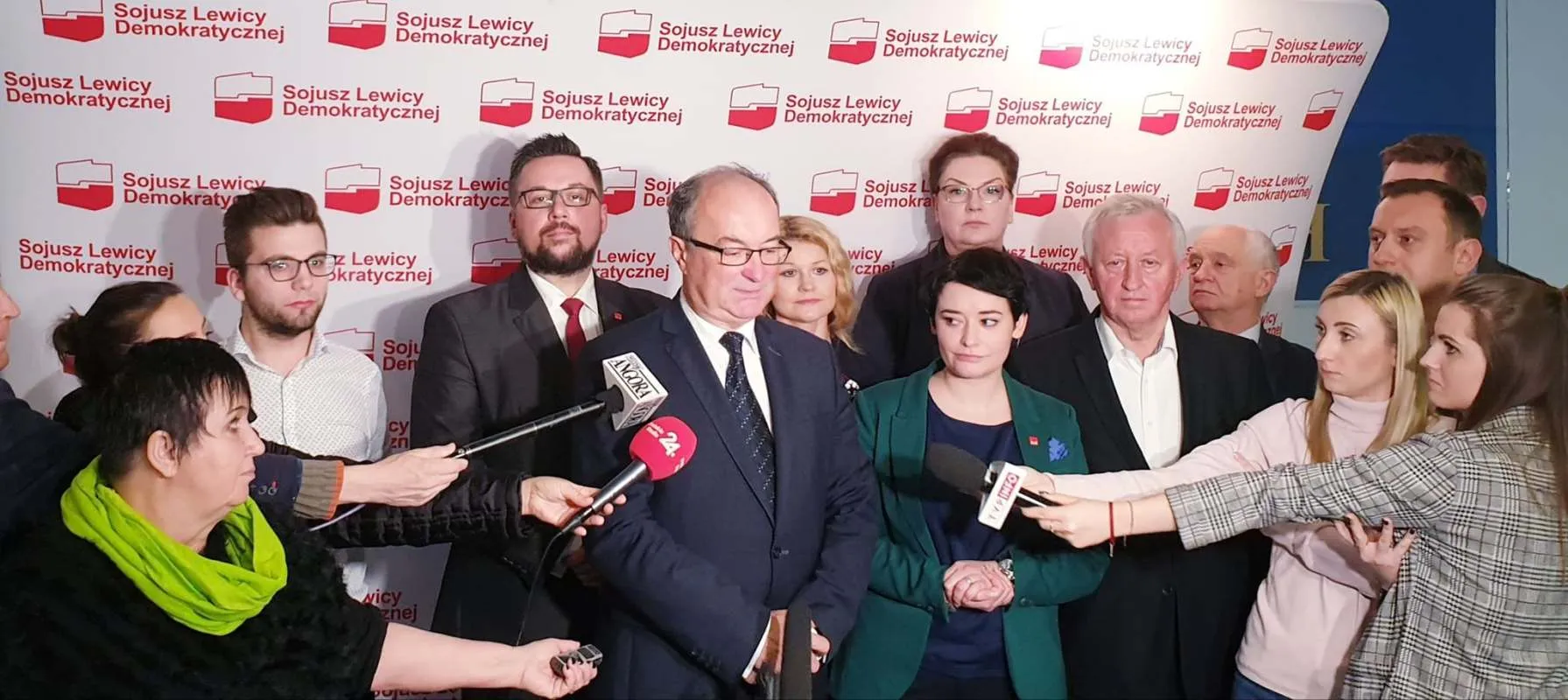 Grupa polityków i dziennikarzy na konferencji prasowej Sojuszu Lewicy Demokratycznej; centralnie przemawia mężczyzna w garniturze, a reporterzy trzymają w jego kierunku mikrofony.