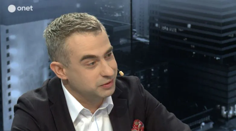 Mężczyzna w garniturze rozmawia w studiu telewizyjnym, z mikrofonem na uchu, na tle miejskiego krajobrazu.