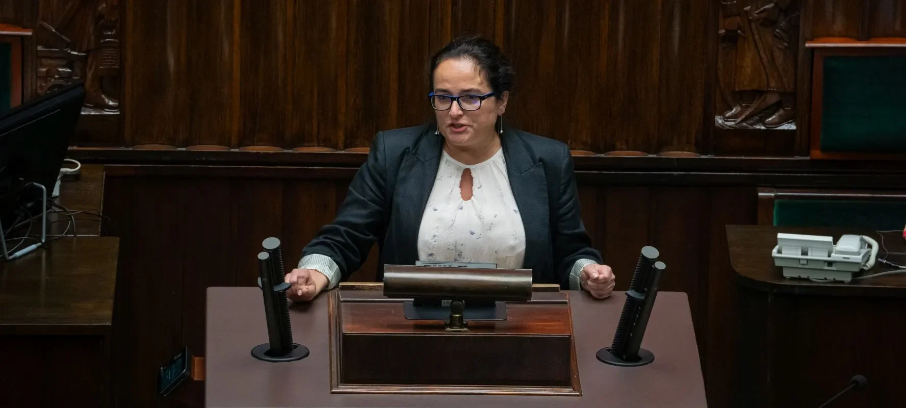 Kobieta w garniturze z niebieskimi okularami stoi za mównicą w sali plenarnej i przemawia.