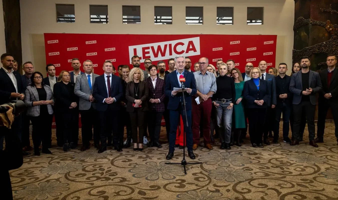 Grupa polityków Nowej Lewicy podczas konferencji prasowej w sprawie umowy koalicyjnej i realizacji programu wyborczego, stojąca przed czerwonym banerem LEWICA, z centralnym mówcą przy mikrofonie.