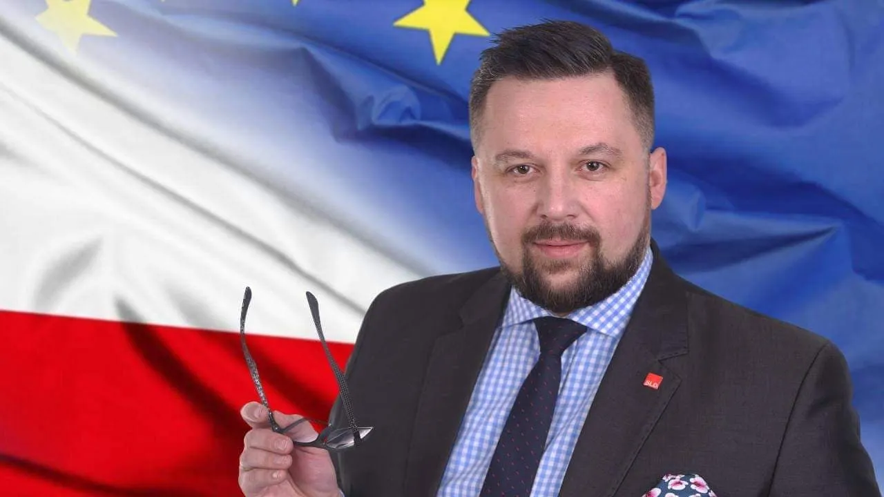 Mężczyzna w garniturze stoi na tle flag Polski i Unii Europejskiej, trzyma w dłoni okulary.