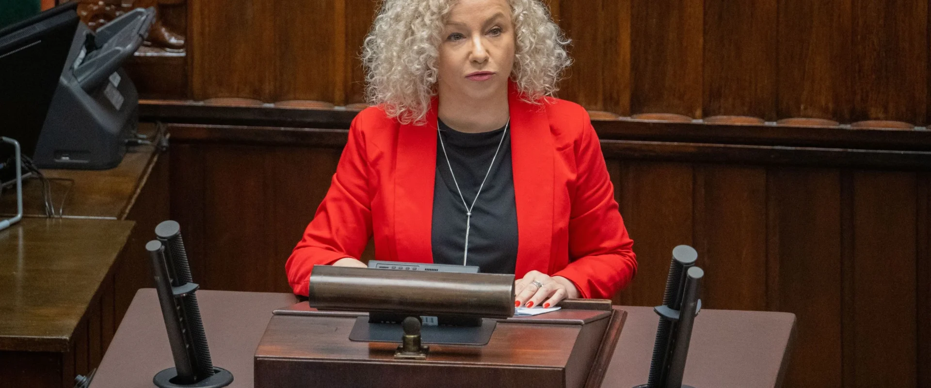 Kobieta o kręconych, siwych włosach w czerwonej marynarce siedzi przy pulpicie w sali obrad.