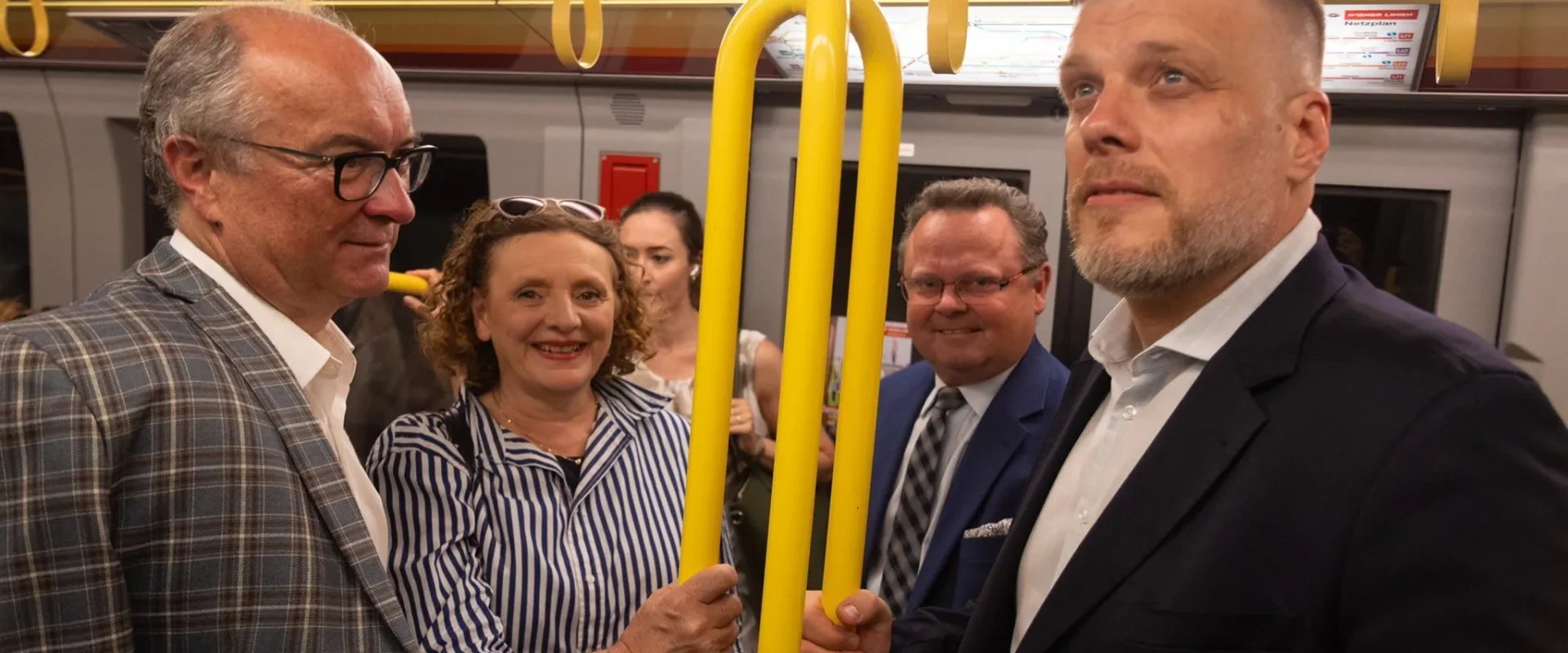 Grupa pasażerów w eleganckich ubraniach stoi w pociągu, trzymając się żółtych uchwytów i rozmawiając.