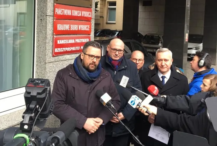 Polityk w ciemnym płaszczu przemawia do mediów podczas konferencji prasowej Koalicji Europejskiej przed wejściem do budynku, otoczony mikrofonami i dziennikarzami.