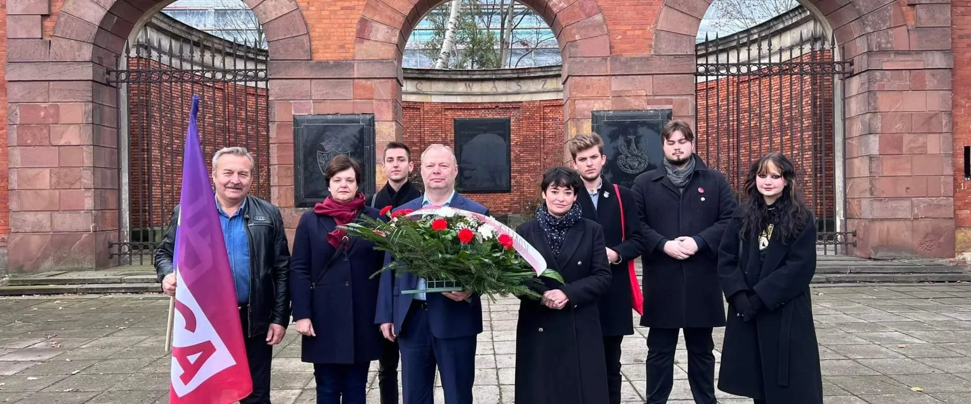 Grupa ludzi stoi przed historyczną bramą, trzymając wieńce i flagi, w celu upamiętnienia ważnej chwili.
