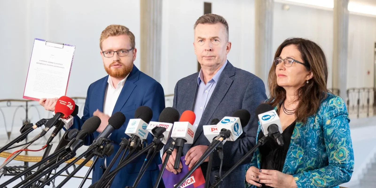 Trzy osoby w garniturach stoją na konferencji prasowej, otoczone licznymi mikrofonami.