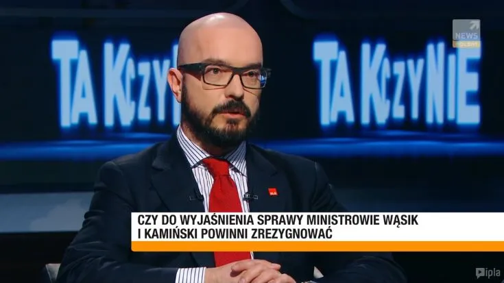 Mężczyzna w okularach i czerwonym krawacie siedzi w garniturze w studiu telewizyjnym, z dużym napisem w tle i paskiem informacyjnym na dole ekranu.