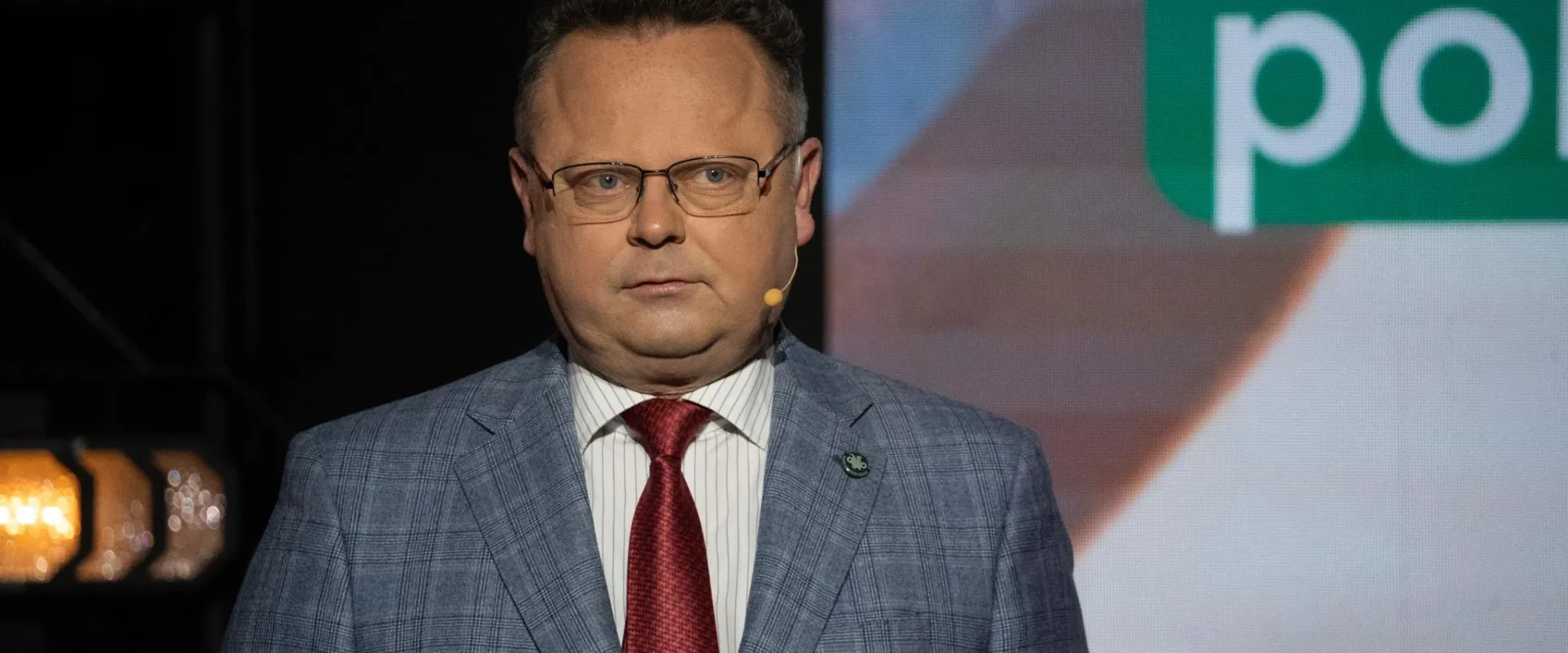Mężczyzna w okularach, garniturze w kratę i czerwonym krawacie przemawia na scenie z mikrofonem przy uchu, a w tle widać duży ekran z napisem.