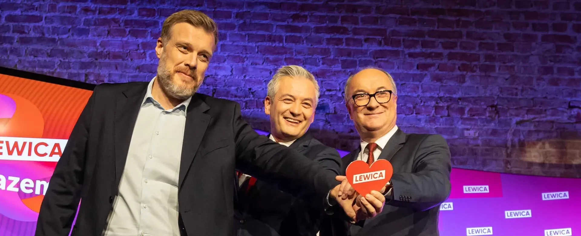 Na zdjęciu trzech mężczyzn pozuje razem, trzymając serce z napisem „LEWICA” w tle kolorowej sceny.