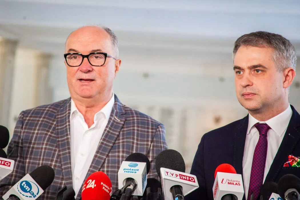 Dwóch mężczyzn w garniturach stoi przy mikrofonach podczas konferencji prasowej.