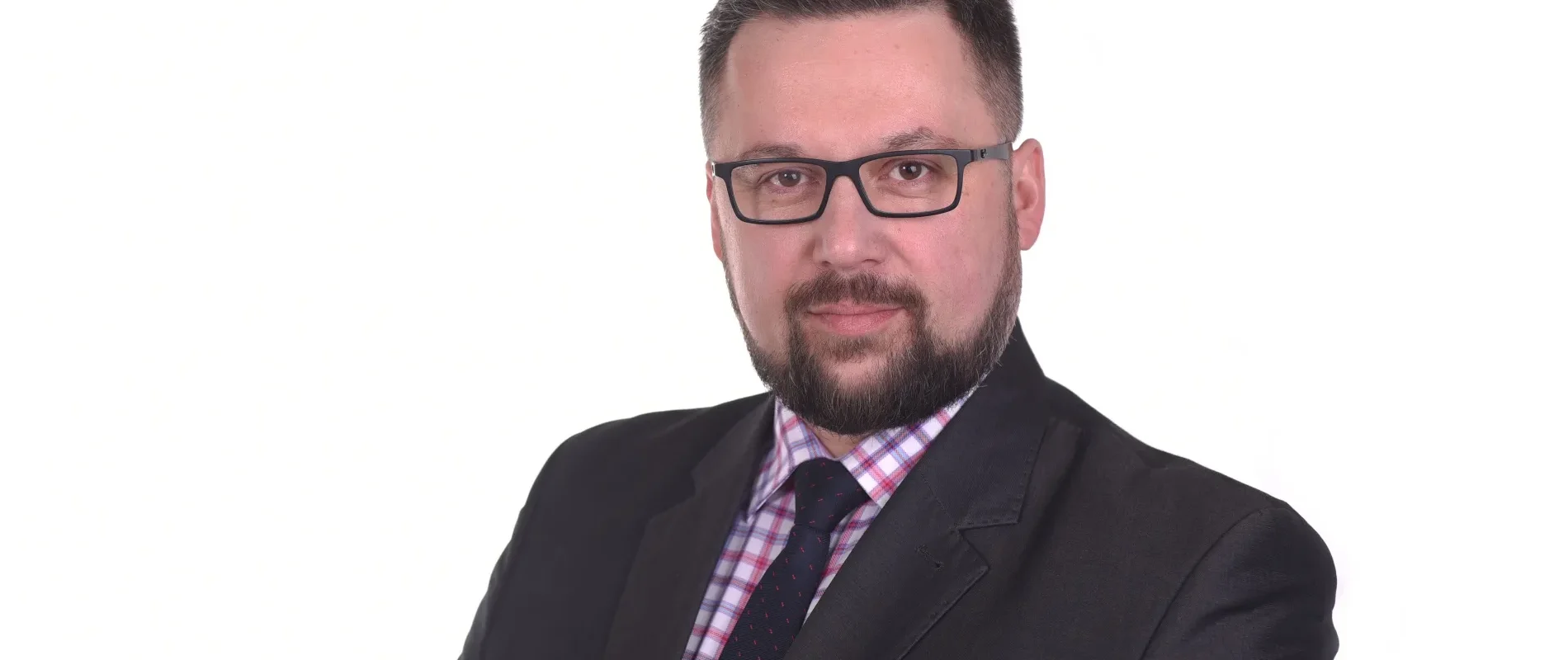 Portret mężczyzny w czarnym garniturze, okularach i z brodą na białym tle.