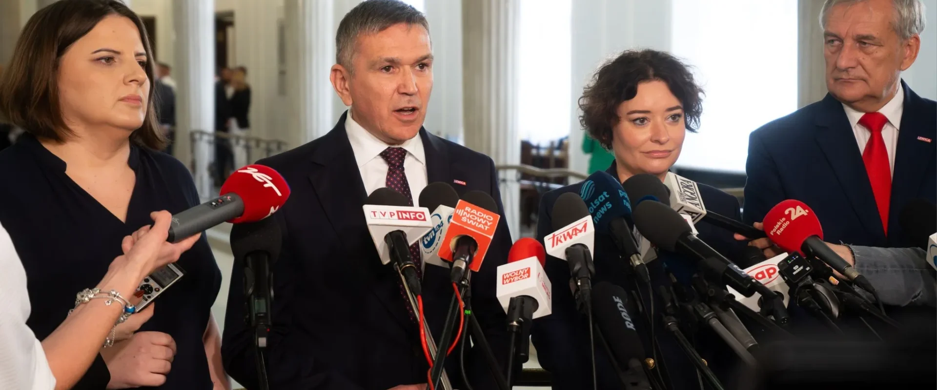 Grupa urzędników i dziennikarzy podczas konferencji prasowej w holu z kolumnami, omawiająca dodatek 2000 zł dla pracowników socjalnych z terenów zalanych, stojąca za licznymi mikrofonami.