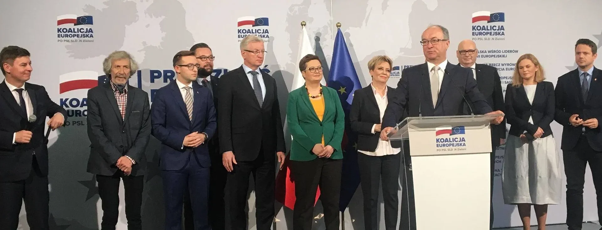 Grupa polityków stoi na scenie przy mównicy z logo Koalicji Europejskiej, w tle widoczne flagi Unii Europejskiej i grafika mapy świata.