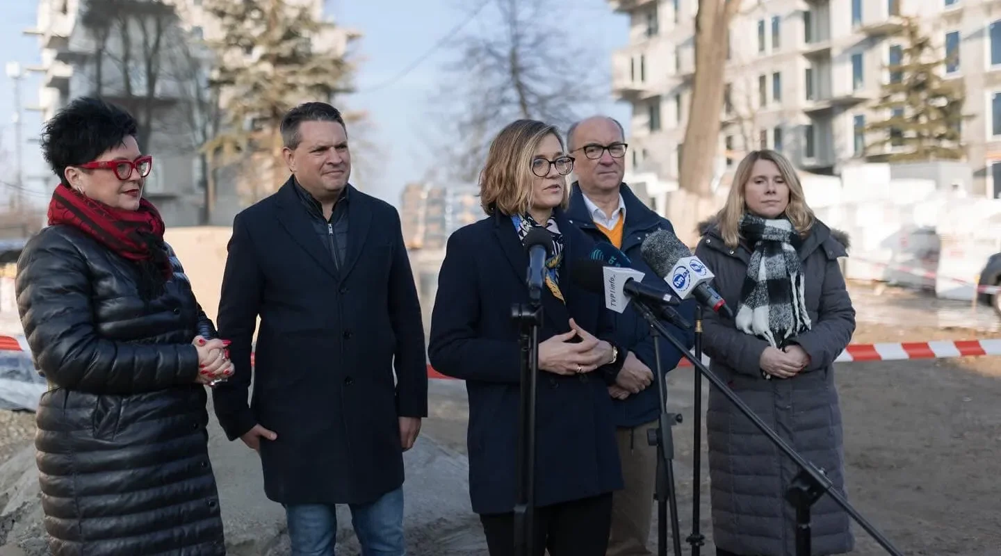 Grupa pięciu osób na konferencji prasowej na placu budowy, w tle blok mieszkalny; kobieta w okularach przemawia do mikrofonów ustawionych przed nimi.