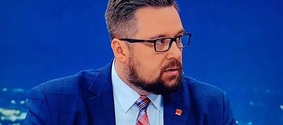 Mężczyzna w granatowym garniturze, jasnoniebieskiej koszuli i czerwonym krawacie, z okularami, na tle niebieskiego tła.