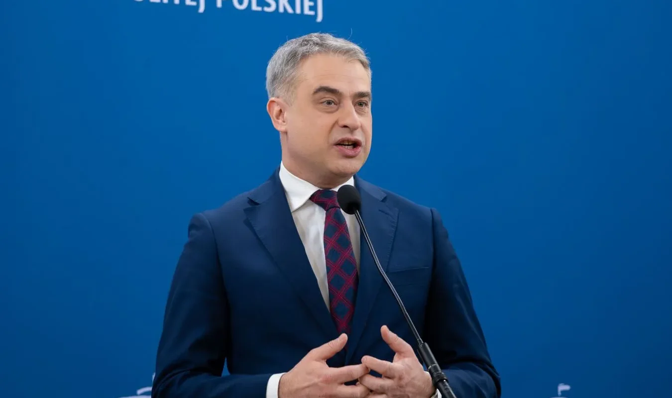 Polityk w granatowym garniturze przemawia do mikrofonu na tle niebieskiej ściany z napisem „Rzeczypospolitej Polskiej”.