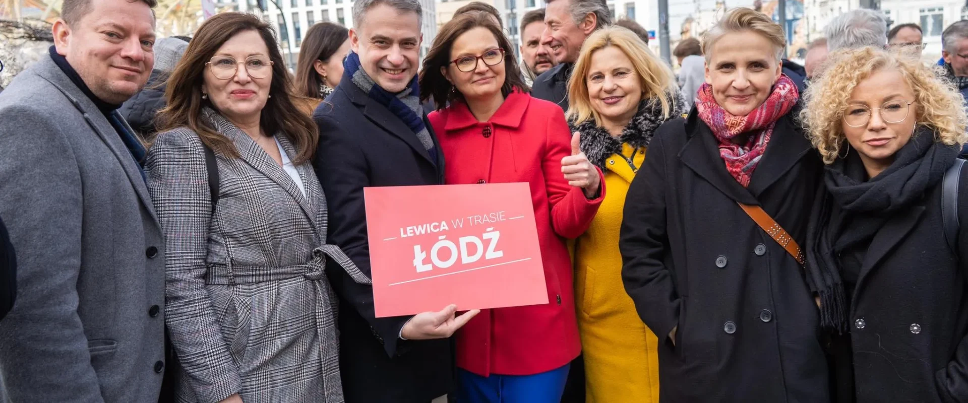 Grupa osób w zimowych ubraniach stoi na placu w Łodzi, trzymając czerwony plakat z napisem „LEWICA W TRASIE ŁÓDŹ”.