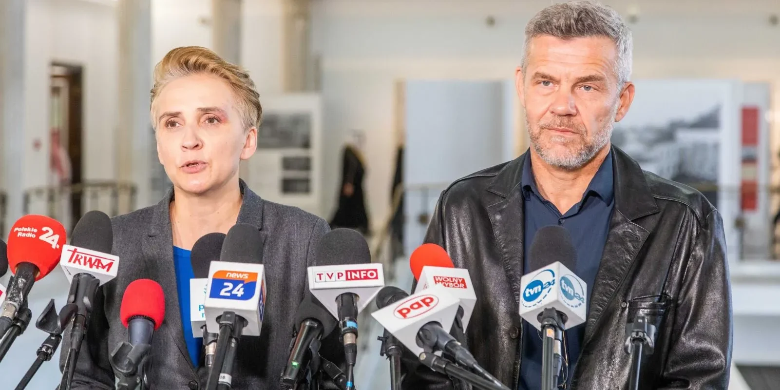 Na zdjęciu widać dwoje mówców znajdujących się przed mikrofonami na konferencji prasowej.