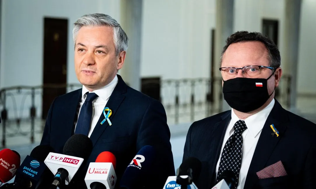 Dwaj mężczyźni w garniturach stoją przed mikrofonami podczas konferencji prasowej w jasnym wnętrzu.