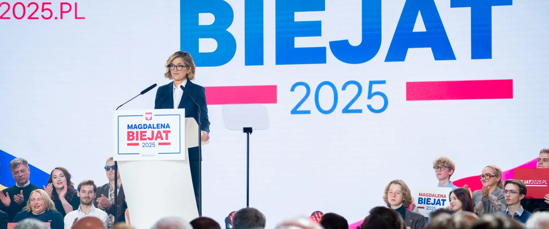 Kobieta w garniturze stoi przy mównicy na scenie podczas wiecu wyborczego, a w tle wyświetlany jest duży ekran z napisem „BIEJAT 2025”.